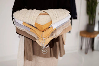 Person holding cozy neutral knit sweaters on wooden hangers in a stylish indoor setting