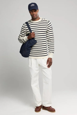 Man in navy striped sweater, white pants, navy cap and bag, brown boat shoes, neutral background