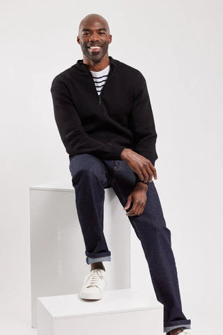 Man in black zip wool cardigan, striped shirt, jeans, and white sneakers sitting on white blocks