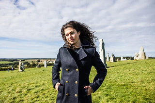 Woman in navy double-breasted pea coat standing outdoors on grassy field with stone monuments