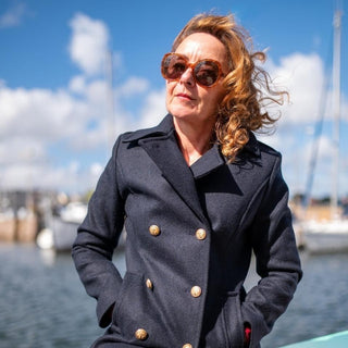 Woman in navy pea coat with gold buttons and sunglasses by a marina on a sunny day