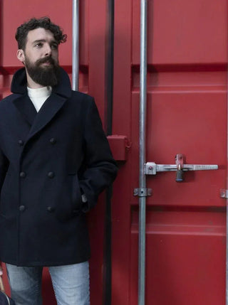 Man in navy wool peacoat and jeans standing by a red industrial container