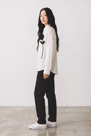 Modèle portant une blouse blanche Le Capitaine D'a Bord à manches longues avec une fleur noire appliquée sur la poitrine