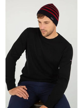 Man wearing a navy and red striped knit beanie and black sweater, sitting indoors