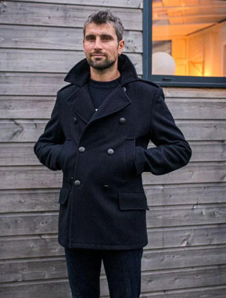 Man wearing a navy wool pea coat with double-breasted buttons standing outside by a wooden wall