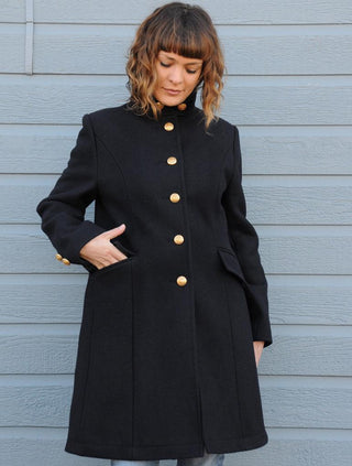 Woman wearing a long navy wool coat with gold buttons, standing by a blue wall.