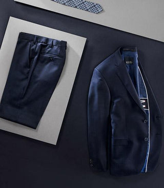 Navy wool suit jacket and pants set laid flat, with a patterned tie nearby on a dark background.